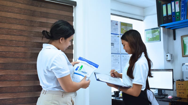 Two Getmeds staff checking a medicine box against a stock list in a pharmacy office